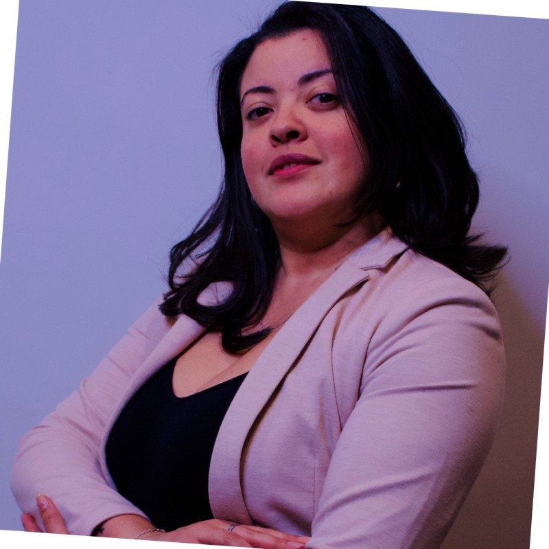 A professional woman wearing a light blazer, with long dark hair and a confident expression, posing with her arms crossed against a plain background.